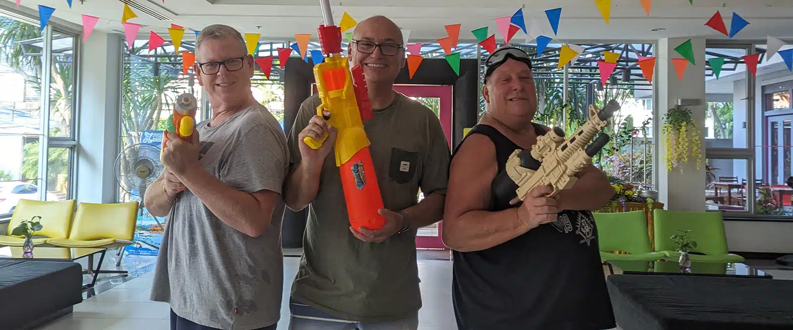 Three men holding water guns, smiling in a lobby decorated with colorful flags.