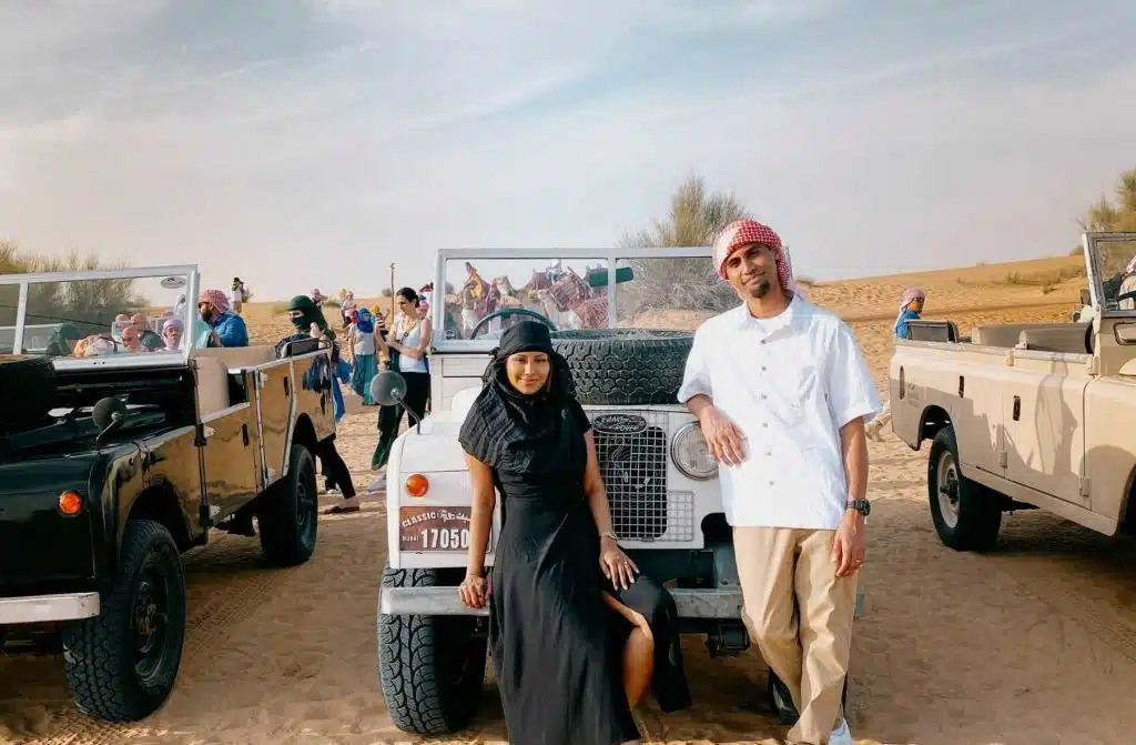 WeWanderlustCo - A man and a woman dressed in traditional arab attire posing in front of a 4x4 vehicle in a desert setting, with people and other vehicles visible in the background.