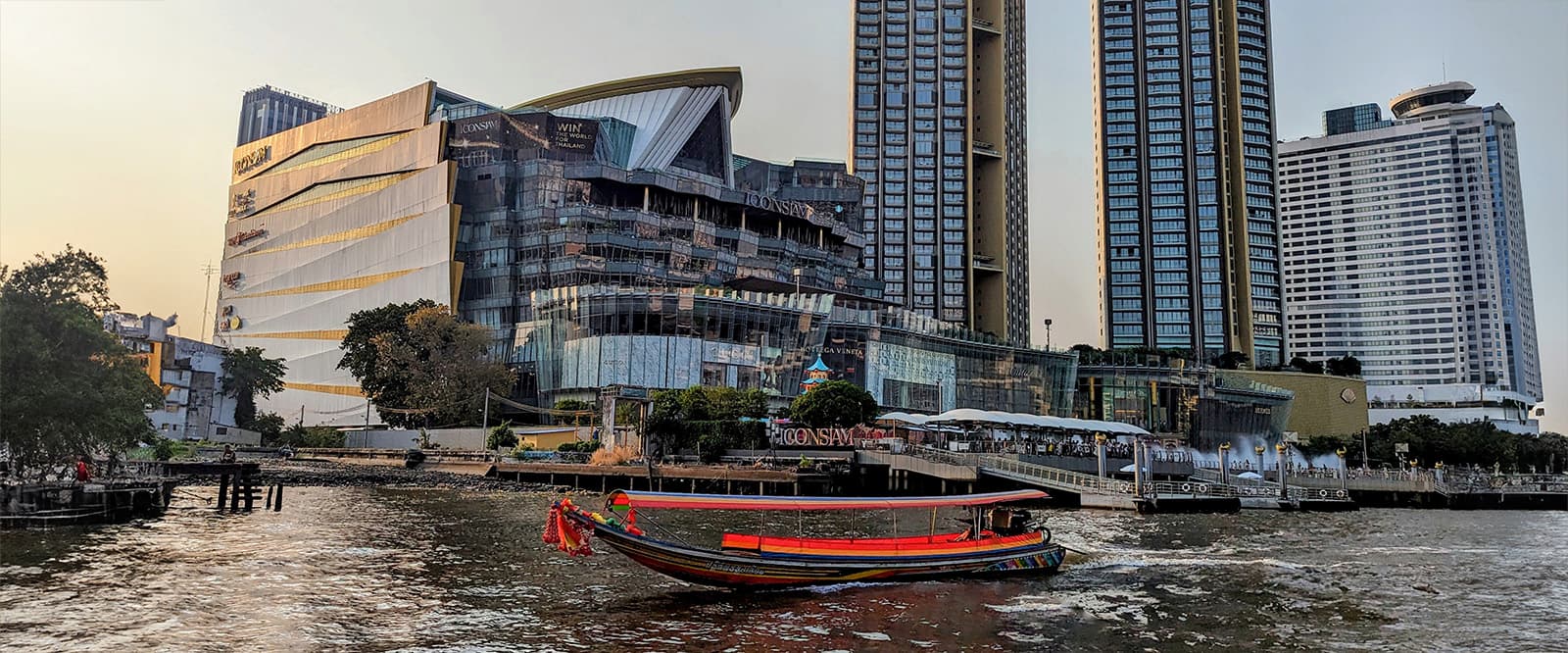 A boat cruising on a Chao Phraya River with modern buildings, such as the Icon Siam along the riverbank under a hazy sky.