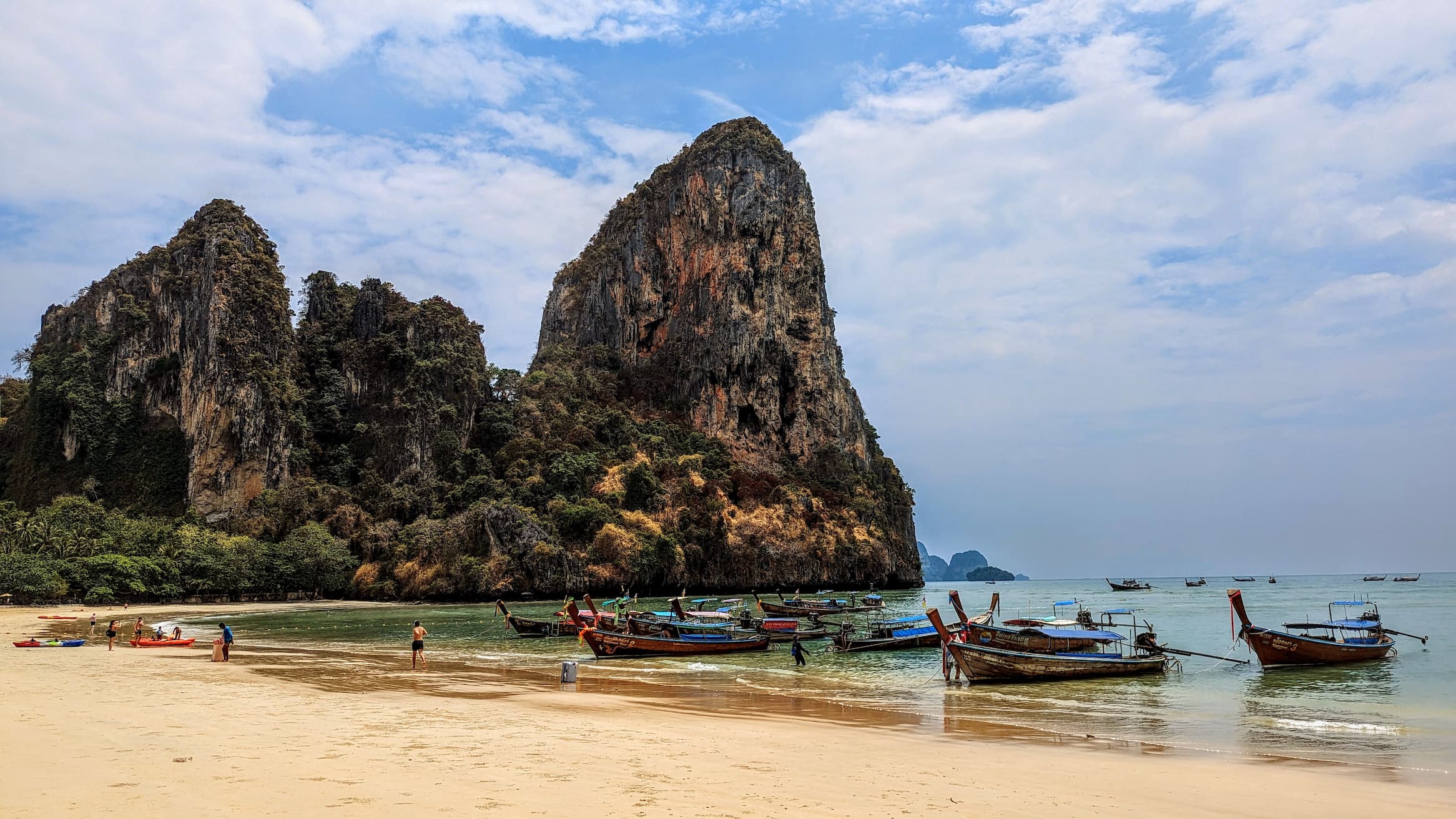 A sandy beach with people and traditional wooden boats in the water; large, rocky cliffs rise in the background against a partly cloudy sky at Railay Bay, Thailand.