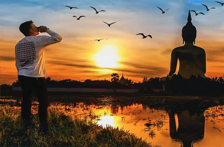 Paddy Doyle standing near a body of water watches a large Buddha statue at sunset, reminiscent of a scene from Paddy Doyle's travels, with birds gracefully flying in the sky.