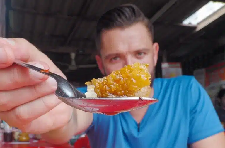 Gary Butler, The Roaming Cook, in a blue shirt holds a spoon with a piece of food close to the camera, capturing the essence of culinary art in an indoor setting.