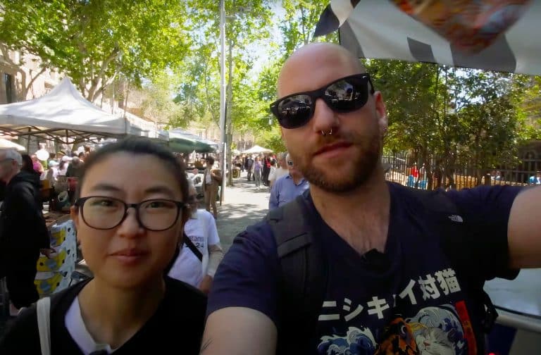Two people from Pasta and Panda travel vloggers are taking a selfie at an outdoor market. Stalls and other visitors are visible in the background on a sunny day.