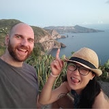 Pasta and Panda travel vloggers strike a fun selfie on a hillside, with dramatic coastal cliffs and the sea in the background. The woman, wearing a hat, playfully sticks out her tongue.