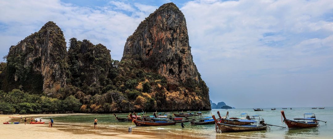 A sandy beach with people and traditional wooden boats in the water; large, rocky cliffs rise in the background against a partly cloudy sky at Railay Bay, Thailand.